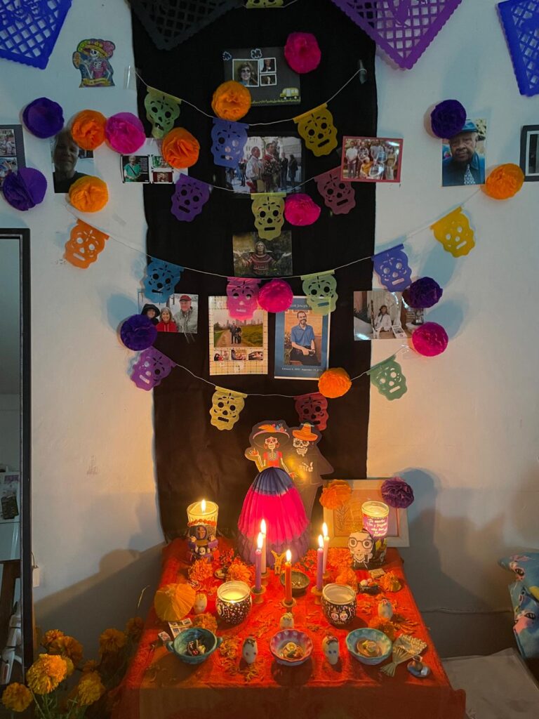 Full Día de Muertos altar decorated with marigolds, candles, colorful paper skulls, and photographs of departed loved ones arranged on a red cloth in a home in Guadalajara.
